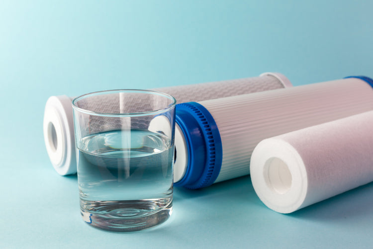 Glass of clear drinking water beside three water filter cartridges on a light blue surface
