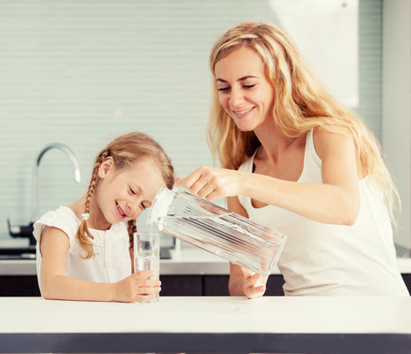A mother pours filtered water from a glass pitcher into her young daughter's glass at a bright kitchen counter