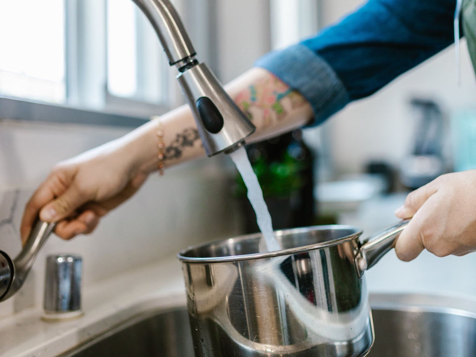 Two hands filling a stainless steel saucepan with tap water from a pull-down kitchen faucet over a stainless sink, bright window light in a home kitchen