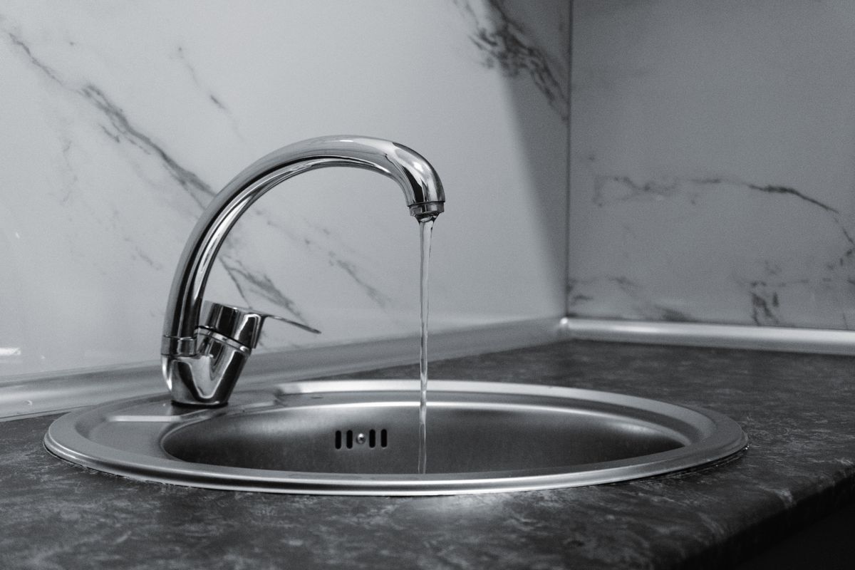 Stainless steel kitchen faucet running a thin stream of water into a round sink, shown in black and white