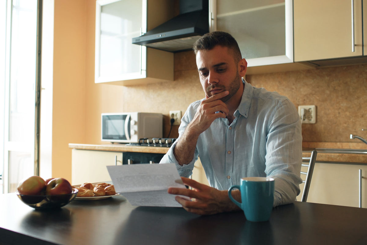 Man reading a printed letter at his kitchen table beside a teal mug, with a kitchen faucet in the background