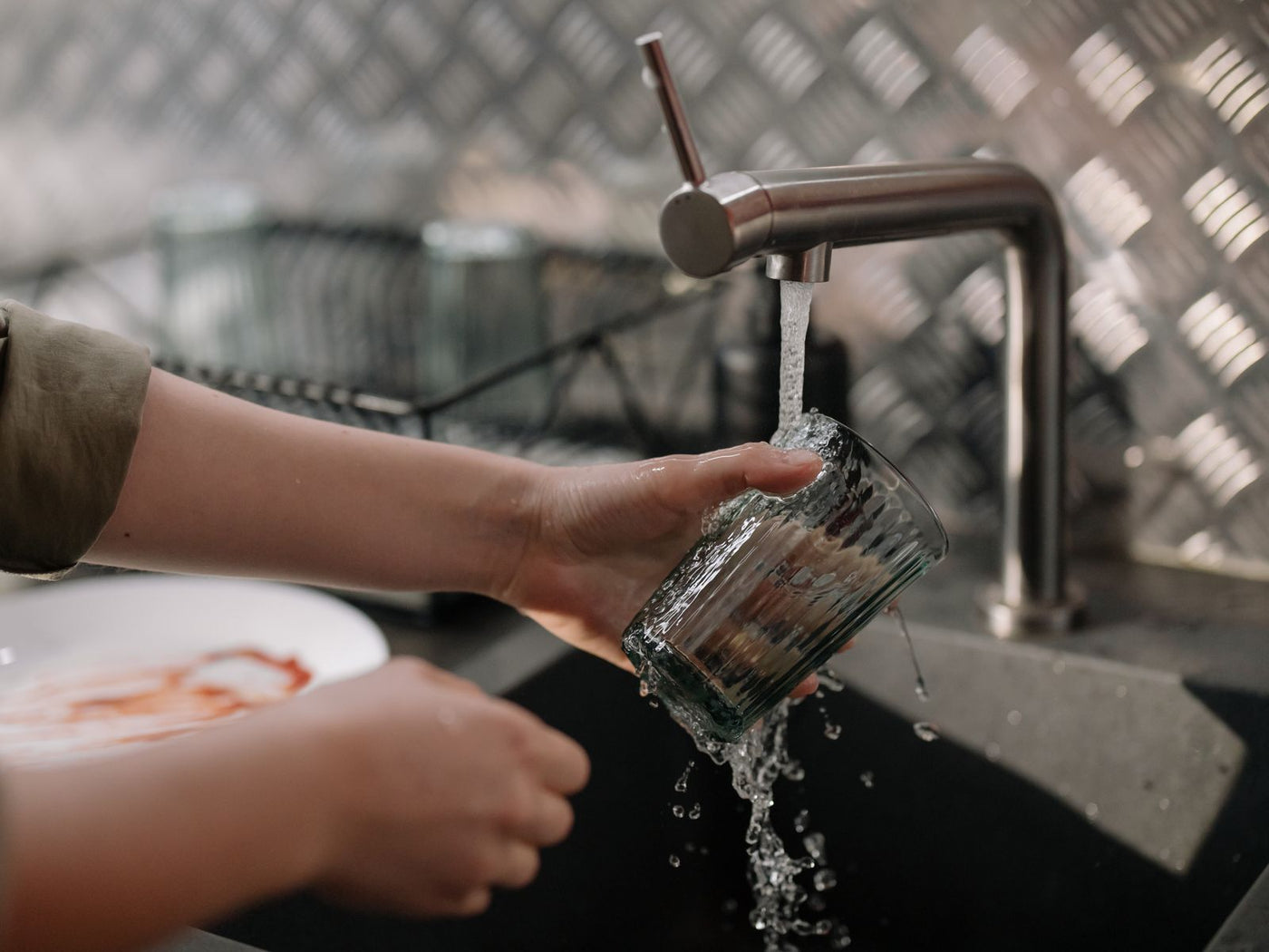 Hands rinsing a drinking glass under running tap water in a stainless kitchen sink