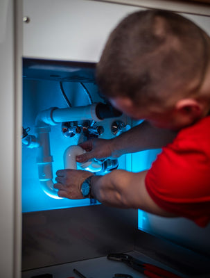Person working in the open cabinet space under a sink, with the P-trap, supply lines, and shutoff valves visible