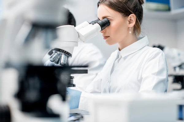 Scientist in a white lab coat examining a sample through a microscope in a bright clinical laboratory